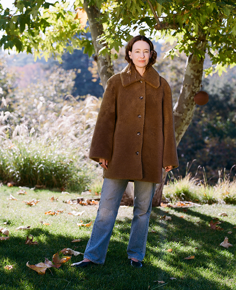 A woman wearing a brown, buttoned winter coat and blue jeans stands on grass under a leafy tree, with sunlight filtering through the branches and a natural, outdoor background.