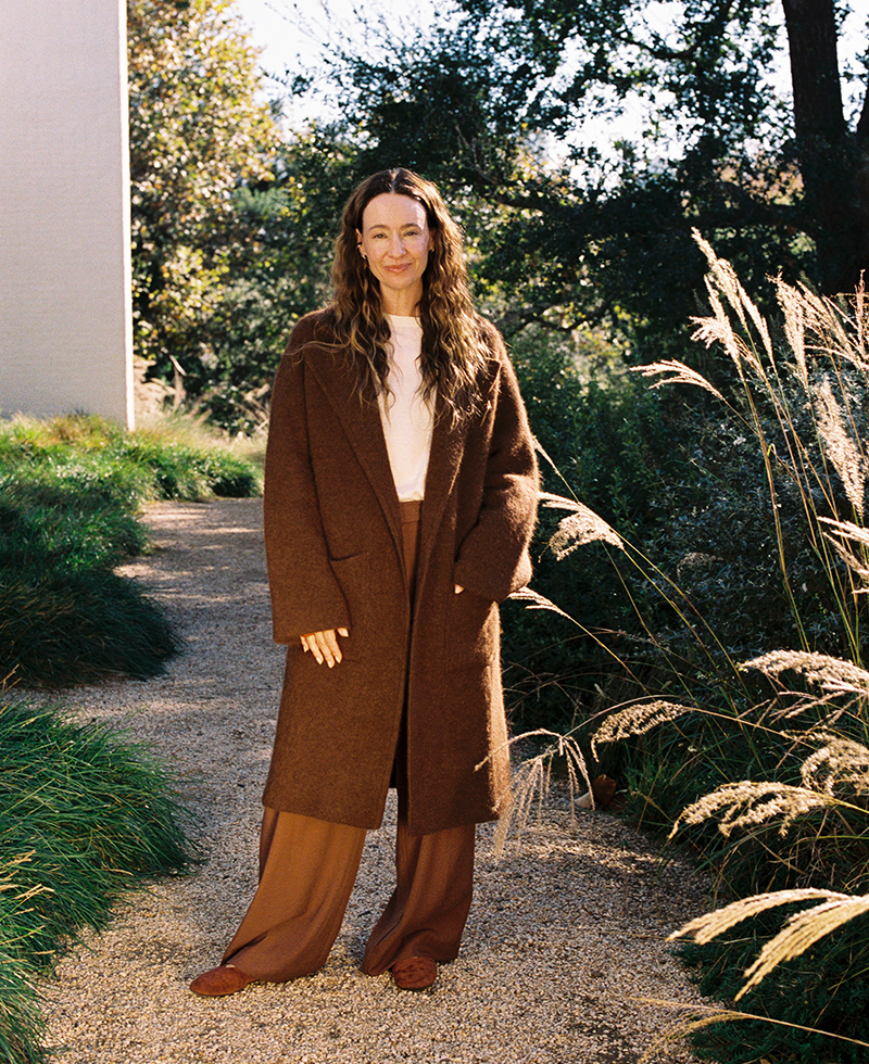 A person with long wavy hair stands on a gravel path outdoors, wearing a stylish brown winter coat, matching brown pants, and shoes. Tall grass and greenery surround the path as sunlight filters softly through the trees.