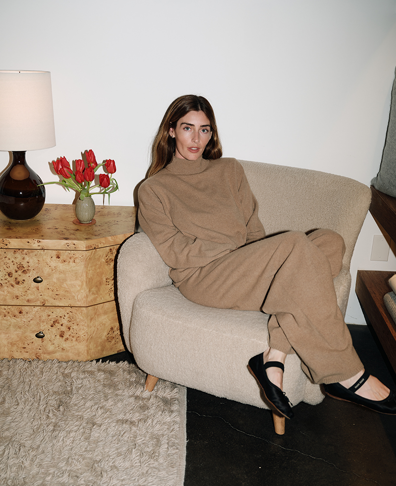 Juliette Labelle, with long brown hair, wears a loose tan outfit and black shoes as she sits on a cream armchair beside a light wood table with a lamp and red tulips. A shaggy beige rug covers part of the dark floor.