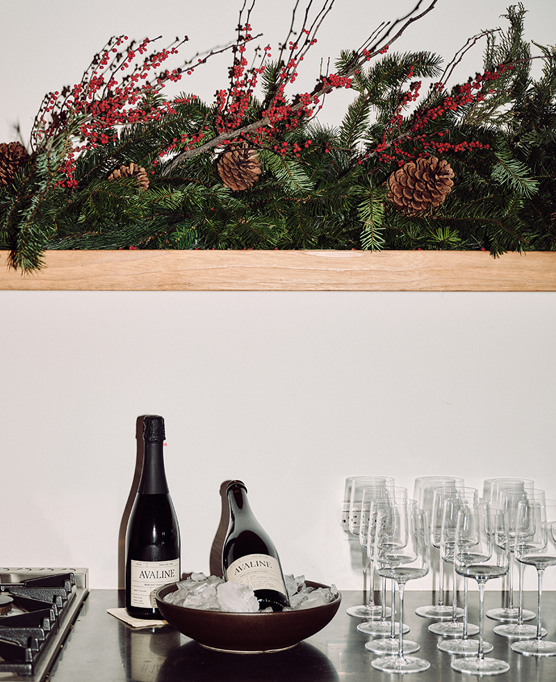 A festive arrangement of pine branches, red berries, and pinecones sits above a shelf. Below, two bottles of Avaline wine by Juliette Labelle rest in a bowl of ice next to several empty wine glasses on a counter.