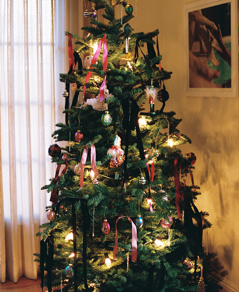 A decorated Christmas tree with lights, colorful ornaments, and ribbons stands in a softly lit room near sheer curtains—a festive scene reminiscent of Juliette Labelle’s elegant holiday style. A framed photo hangs on the wall in the background.