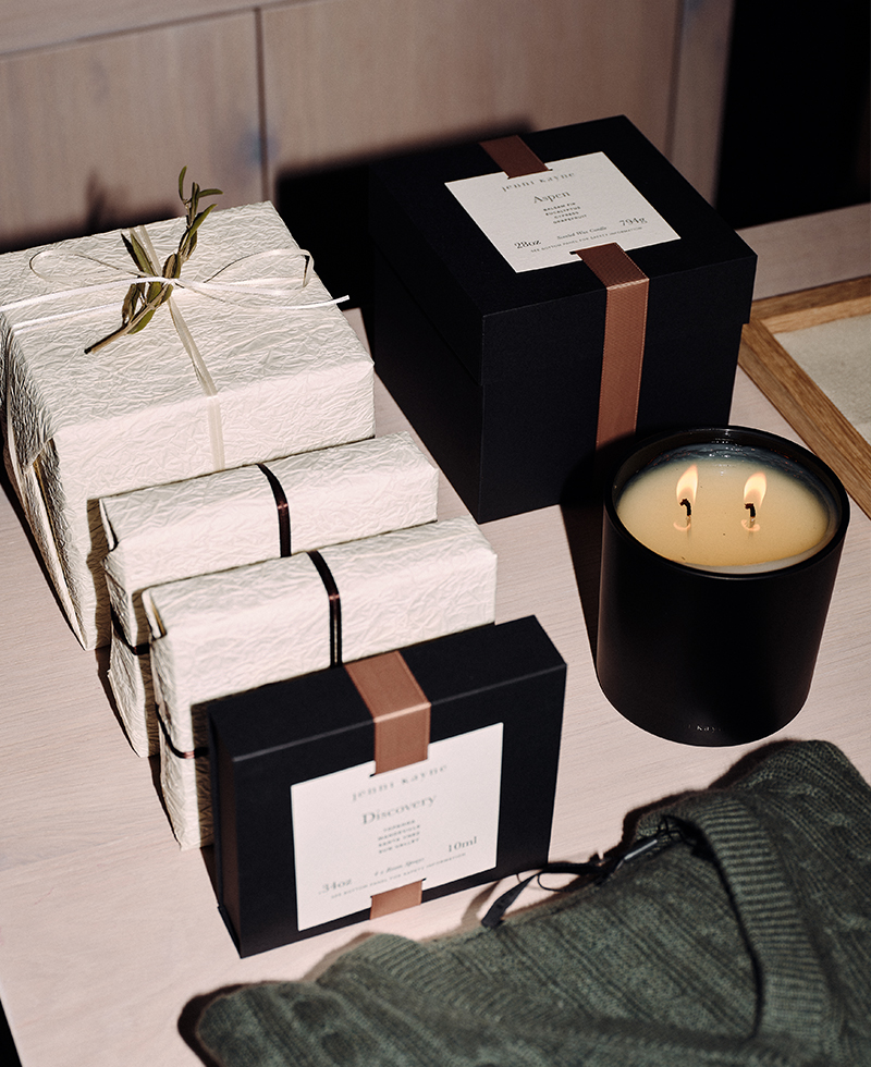 A collection of elegantly wrapped gift boxes, some white with black and gold ribbons and others black with beige ribbons, are arranged on a table next to a lit double-wick candle and a green knit fabric, evoking Juliette Labelle's timeless style.