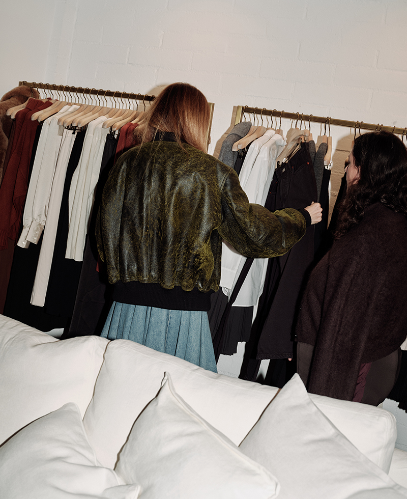Two women stand in front of a clothing rack with various jackets, shirts, and pants, examining the garments. A white couch is in the foreground, and the stylish setting evokes a Juliette Labelle boutique or showroom.