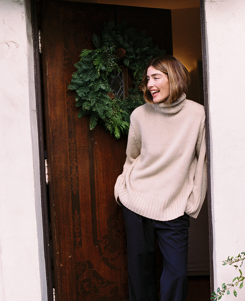 Juliette Labelle stands in a doorway, smiling in a beige turtleneck sweater and dark pants. A green holiday wreath hangs on the wooden door behind her.