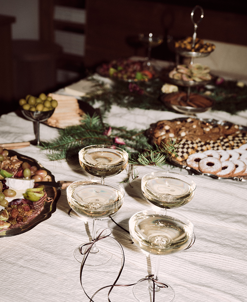 A festive table set with four glasses of sparkling wine, surrounded by holiday treats, cookies, olives, fruit, and greenery on a white tablecloth—elegantly styled in the spirit of Juliette Labelle.