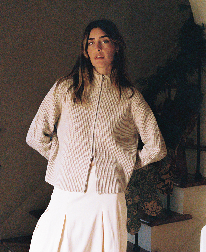 Juliette Labelle, with her long brown hair, stands indoors in front of stairs, wearing a light beige ribbed zip-up sweater and a cream pleated skirt, with her hands behind her back.