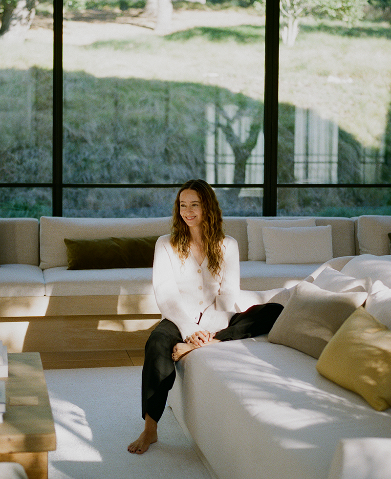 jenni sitting in her living room on a beige couch wearing black trousers and an ivory cardigan
