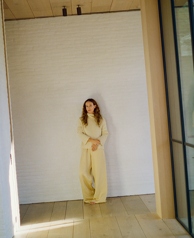 jenni wearing a cashmere turtleneck in yellow and yellow pants leaning against an ivory brick wall