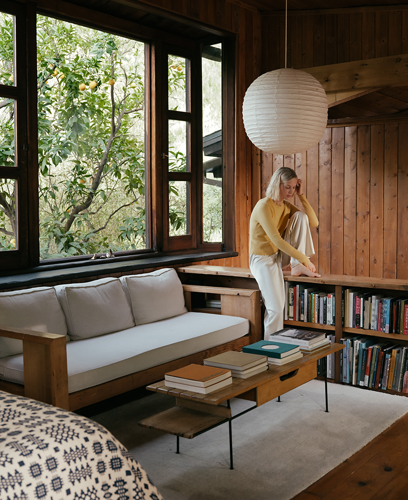 kate parfet wearing a yellow sweater and ivory pants sitting on a ledge in her bedroom with brown wood paneled walls