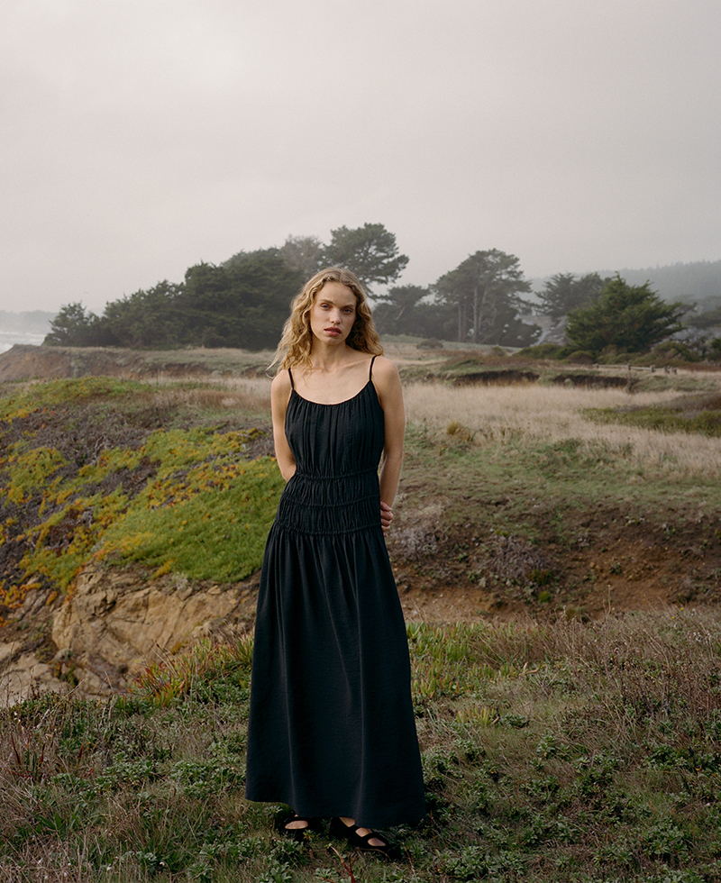 a woman in a black long dress standing on a cliff at sea ranch